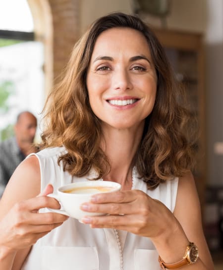 A woman with dark hair sits inside and smiles at the camera while holding up a coffee.