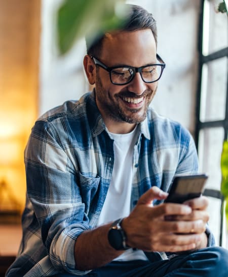 A man with dark hair and glasses sits on a stool inside and smiles while looking down at his phone.